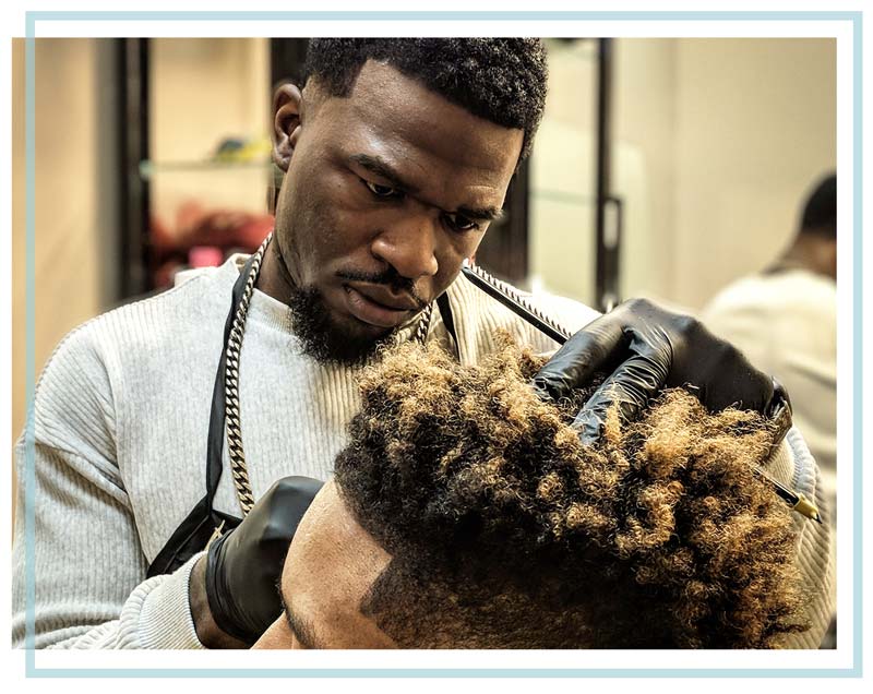 A Black male barber wearing a mask gives a haircut to a Black male patron.