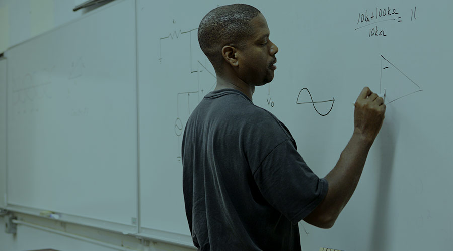 A Black male college student writes at a classroom whiteboard