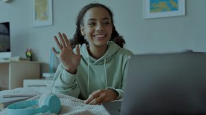 An African American business woman sits at a desk in front of a computer. She has a slight smile and her hand is lifted as if in response to a question