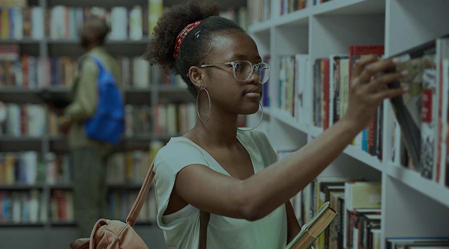 The image shows a serious Black student with glasses in a well-lit library, taking a book off of a shelf