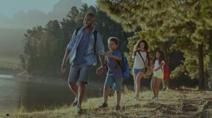 An image of a Black family hiking a trail next to a lake shows the father taking lead. They are smiling and relaxed