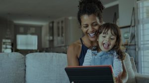 The picture shows a young Black mom holding her toddler in her lap on a gray cloth sofa. They are looking at a laptop monitor and laughing joyfully together