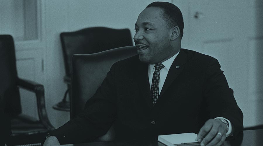 An image shows the great Reverend Dr. Martin Luther King Jr. facing to the left side. King is seated at a desk and holding a paper
