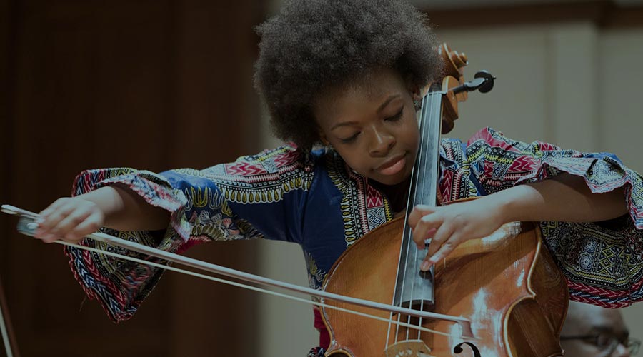 In the image a young Black musician plays the cello. Their hair is natural and they are wearing a multicolored tunic