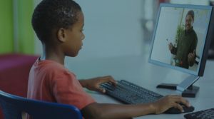 In a photograph, an elementary-school aged Black child sits at a desk with a computer observing a lesson on a monitor using the keyboard with confidence