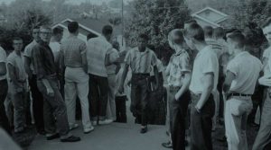 A Black high school student walks up steps onto a school campus with books in his hand surrounded by white students who turn to stare at him