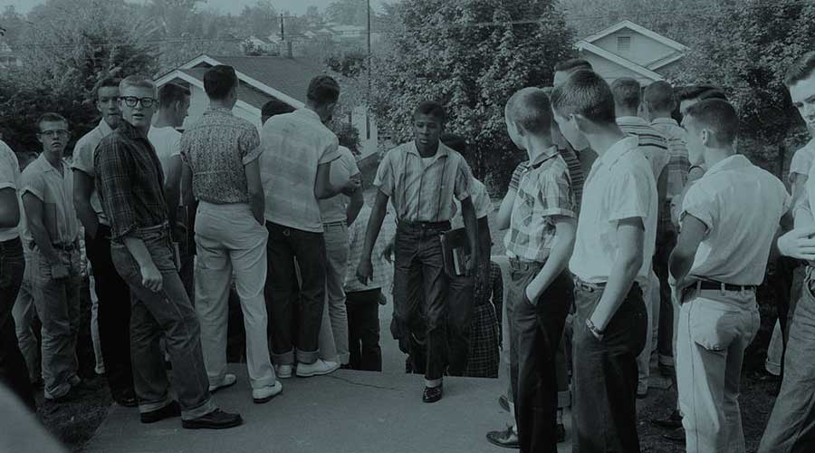 A Black high school student walks up steps onto a school campus with books in his hand surrounded by white students who turn to stare at him