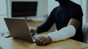 A young Black man sits at a desk in front of a laptop. His arm, also resting on the table, is bandaged