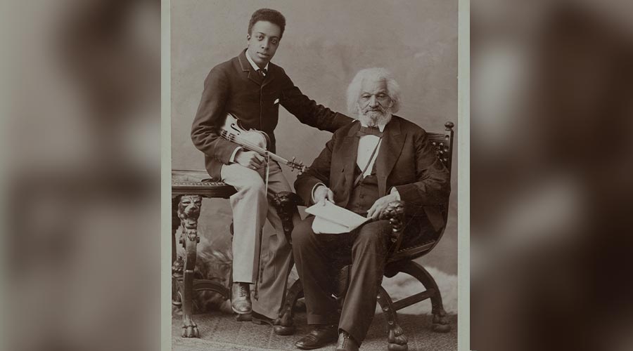 A young Black man holds a violin and sits on a table behind author Frederick Douglas who is seated on a chair