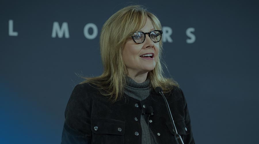 Mary T. Barra, the Chair and CEO of General Motors, wears glasses and speaks at a microphone to an audience off camera