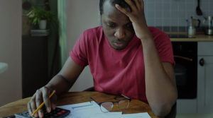 A young Black man is seated at a desk with his head in his hand as he looks at a number of bills