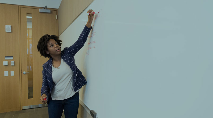A young Black woman stands in a suit jacket while giving a presentation at a white board.