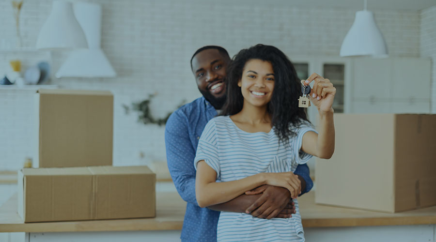 A young Black couple stand holding up a key chain and smile in front of moving boxes
