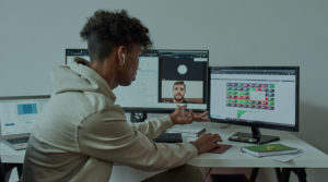 Sitting at a desk with multiple screens open, a young black man, talks on Zoom while working