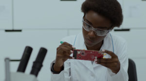 A young black engineer wears protective glasses and a white shirt in a lab while working on a small circuit board