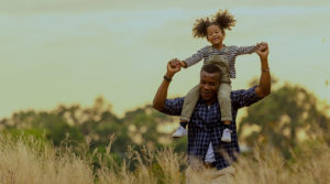 A young Black father walks laughing through a field of tall grass with his daughter on his shoulders