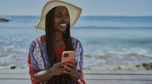 An African American woman in a sun hat with her back to the ocean takes a selfie