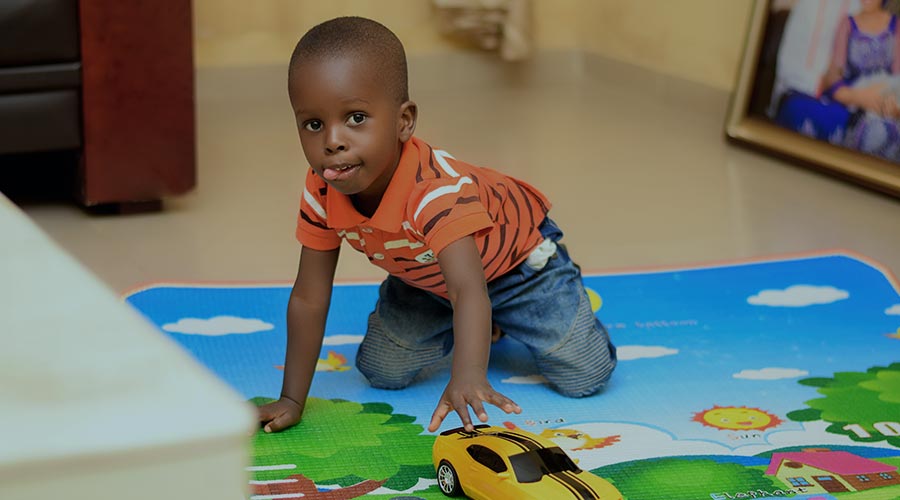 A Black kindergarten-aged child in an orange shirt pushes a yellow sports car on a mat