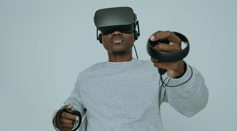 A young Black man in a white long sleeve shirt wears a virtual reality visor and uses hand controls