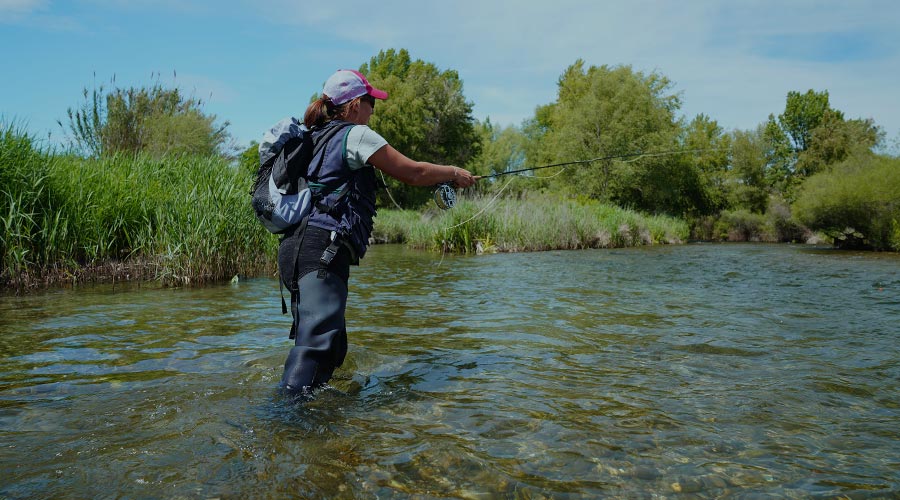 A woman stands in a stream wearing fishing gear and holds a fishing rod as she casts the line to find a fish