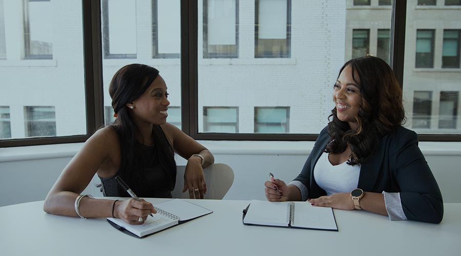 Two Black women executives sit at a conference table in a sunlight room with notebooks open to brainstorm ideas