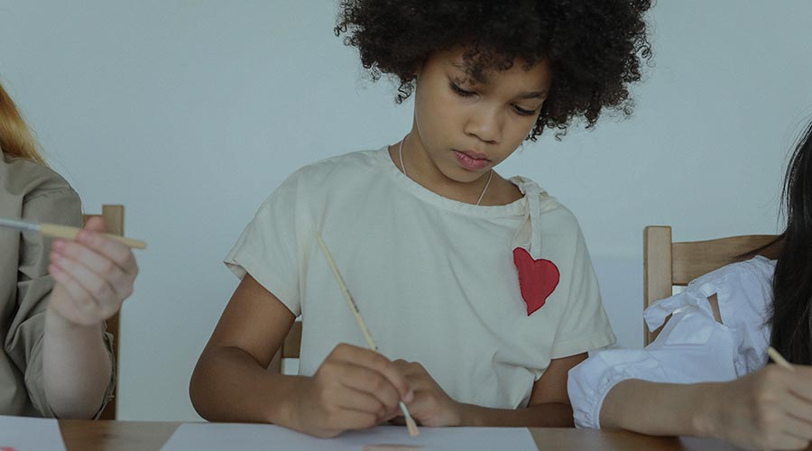 A young Black student with a heart embroidered on her shirt holds a paint brush over to paper in a class with other children