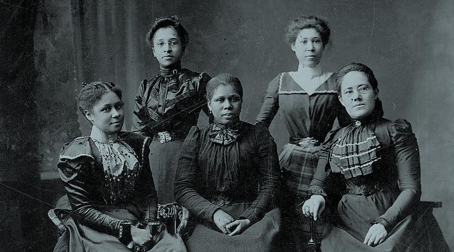 Five Black women grouped in a black and white photograph from 1900 wear dresses and embellishments from the era