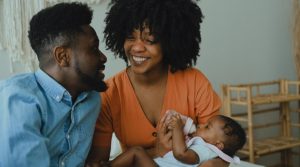 A Black husband and wife smile at each other. She is holding an infant