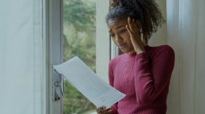 Black woman in her late twenty's wearing a pink sweater holds a bill in one hand and places her hand to her temple