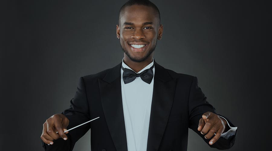 A Black classical music composer stands wearing a black and white tuxedo with his arms in position to conduct an orchestra