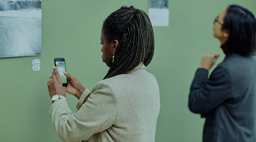 A Black woman in a light suit jacket uses her smartphone and QR code to navigate a digital tour in a museum