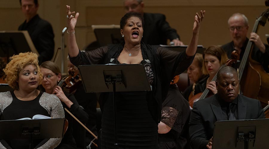 A Black woman in a black gown raises her arms to sing on stage at Carnegie Hall