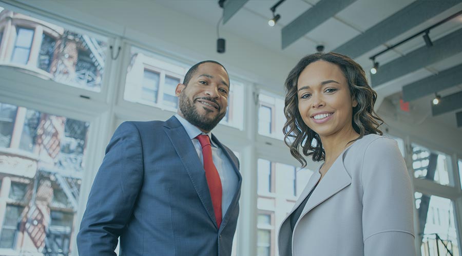 Two African American adults smile while wearing suits and standing in an office