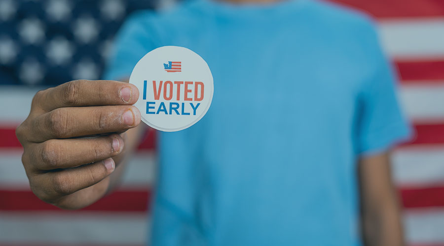 A young person seen from the neck down holds out a sticker that says: "I Voted Early."