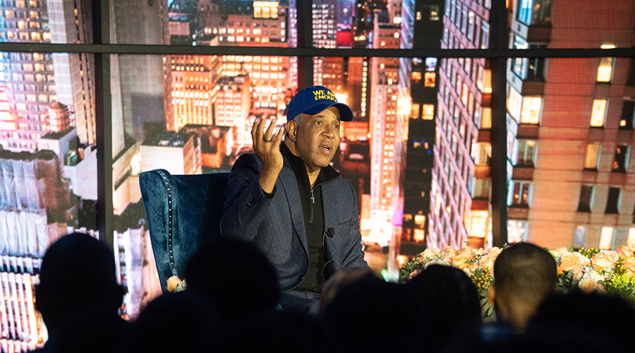 Robert F. Smith sitting on a stage in a baseball cap, blazer in front of an audience