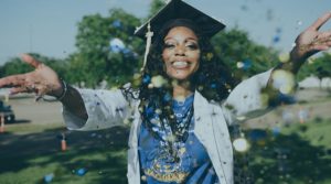 A young Black woman wearing a graduation cap, white lab coat and a stethoscope celebrates by throwing her hands out and up