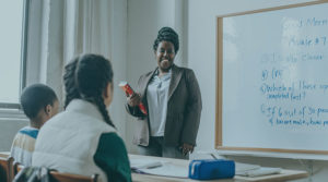 An African American teacher with natural braided hair stands in front of Black elementary school students