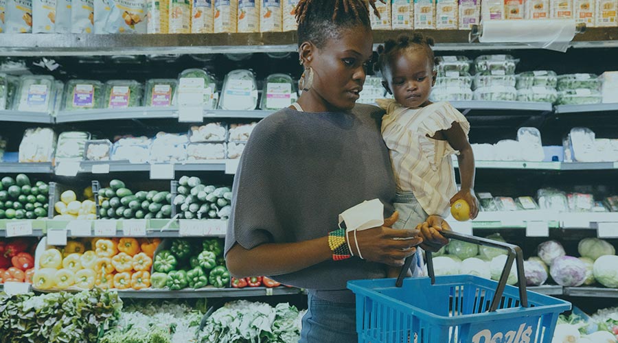 A young Black mother holds her toddler and a shopping basket in the produce section of a grocery store