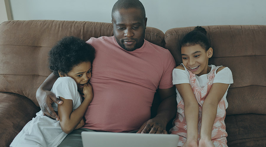 A black man sitting on the couch with two children looking at a laptop screen