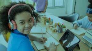 A young Black girl sits at a table with headphones on while writing in a notebook.