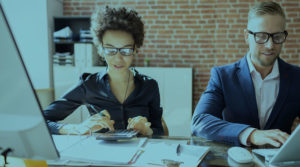 A Black woman sits at a desk using a calculator alongside a man typing on a laptop.