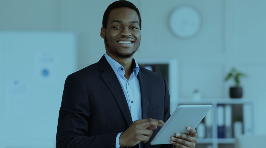 A young African American man wearing a suit holds a digital tablet while smiling at the viewer.