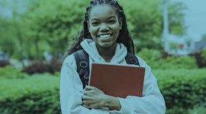 A photo of a young Black girl smiling while she holds a notebook in her hands.