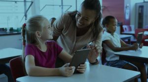 A Black teacher smiles and leans down to help a young girl while she sits at a desk.