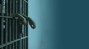 A photo of a jail cell while a inmate's hands hang outside of the bars.