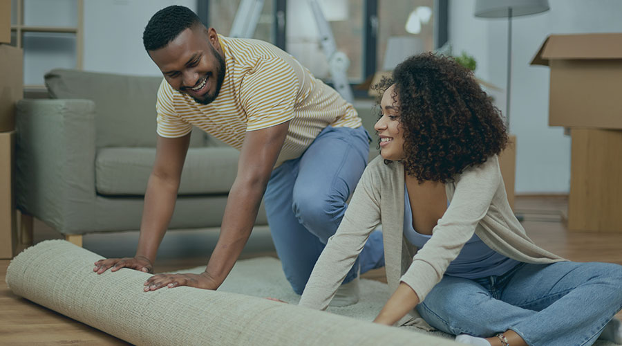 An image of two young African American people on a wood floor rolling up an area rug.