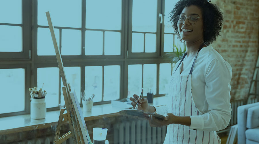 A young Black woman smiles with an easel in front of her as she paints a work of art.