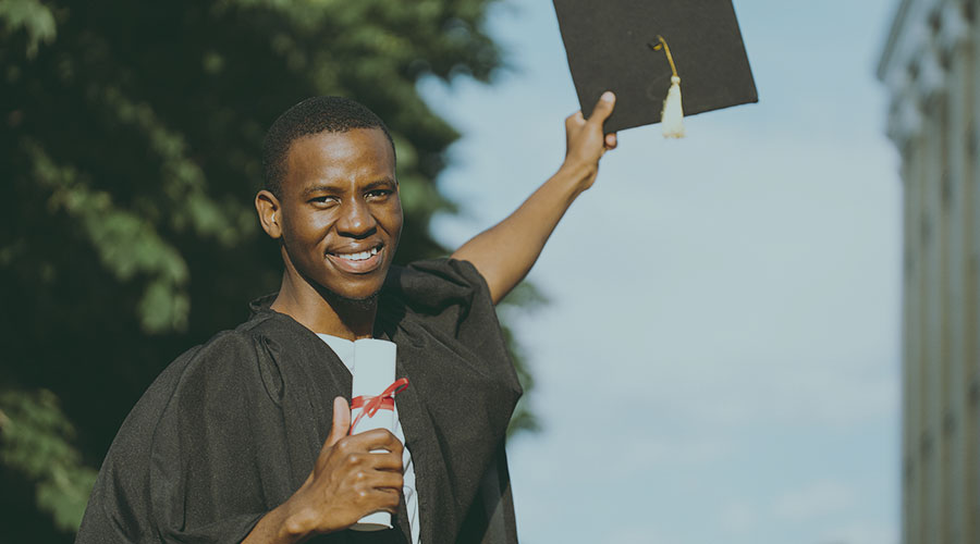 Image of an African American man wearing a black graduation gown and holding a graduation cap and diploma