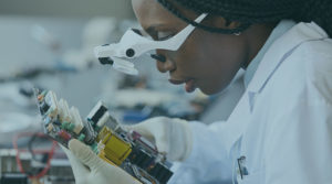 Image of a Black woman in a white lab coat, wearing white magnifying glasses and holding a piece of technology.