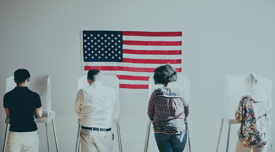 Four people standing at voting machines in front of an American flag.